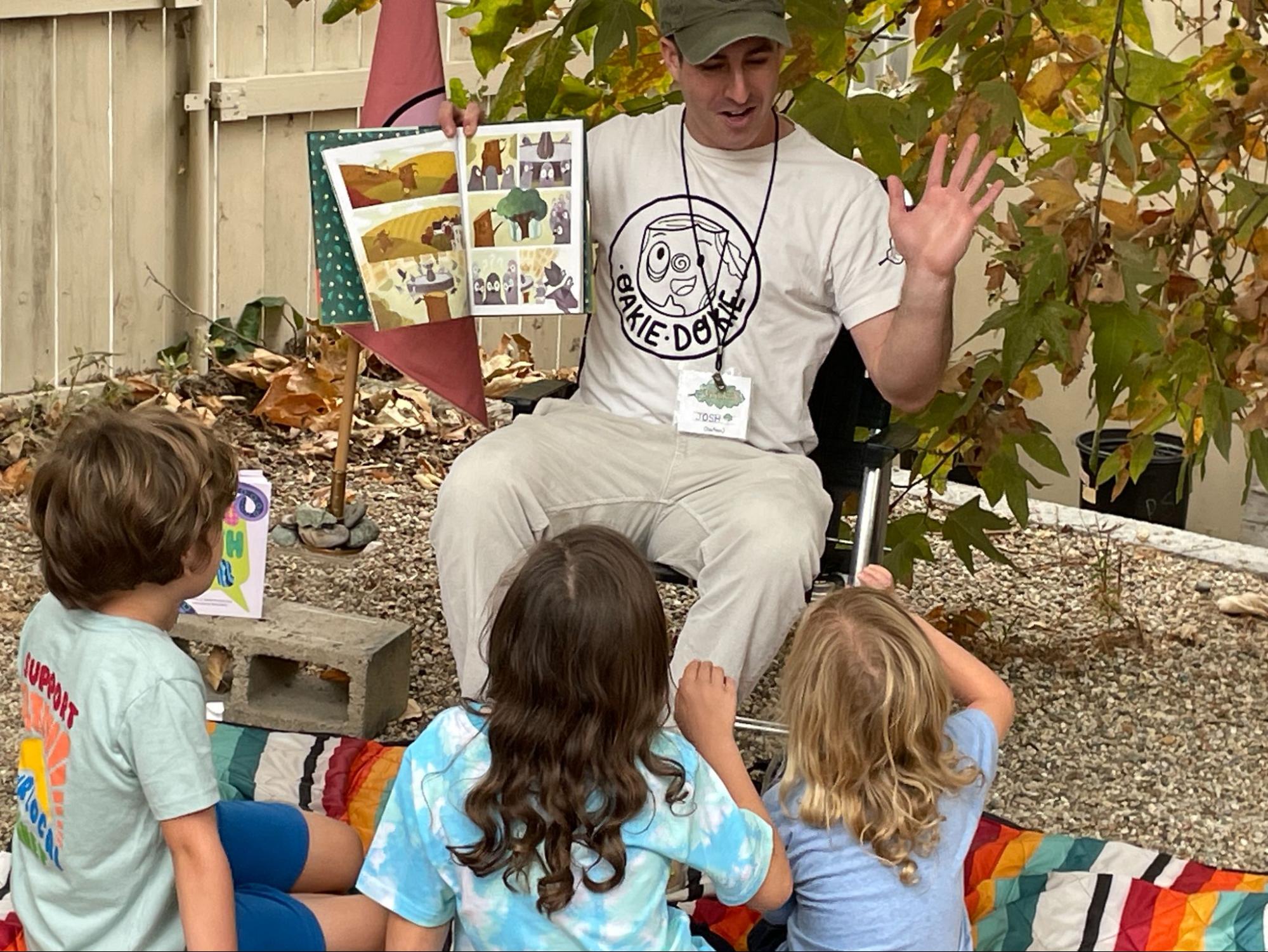 Josh Oaktree seated outdoors reading to three young children gathered on a blanket, holding up an illustrated page from one of his books