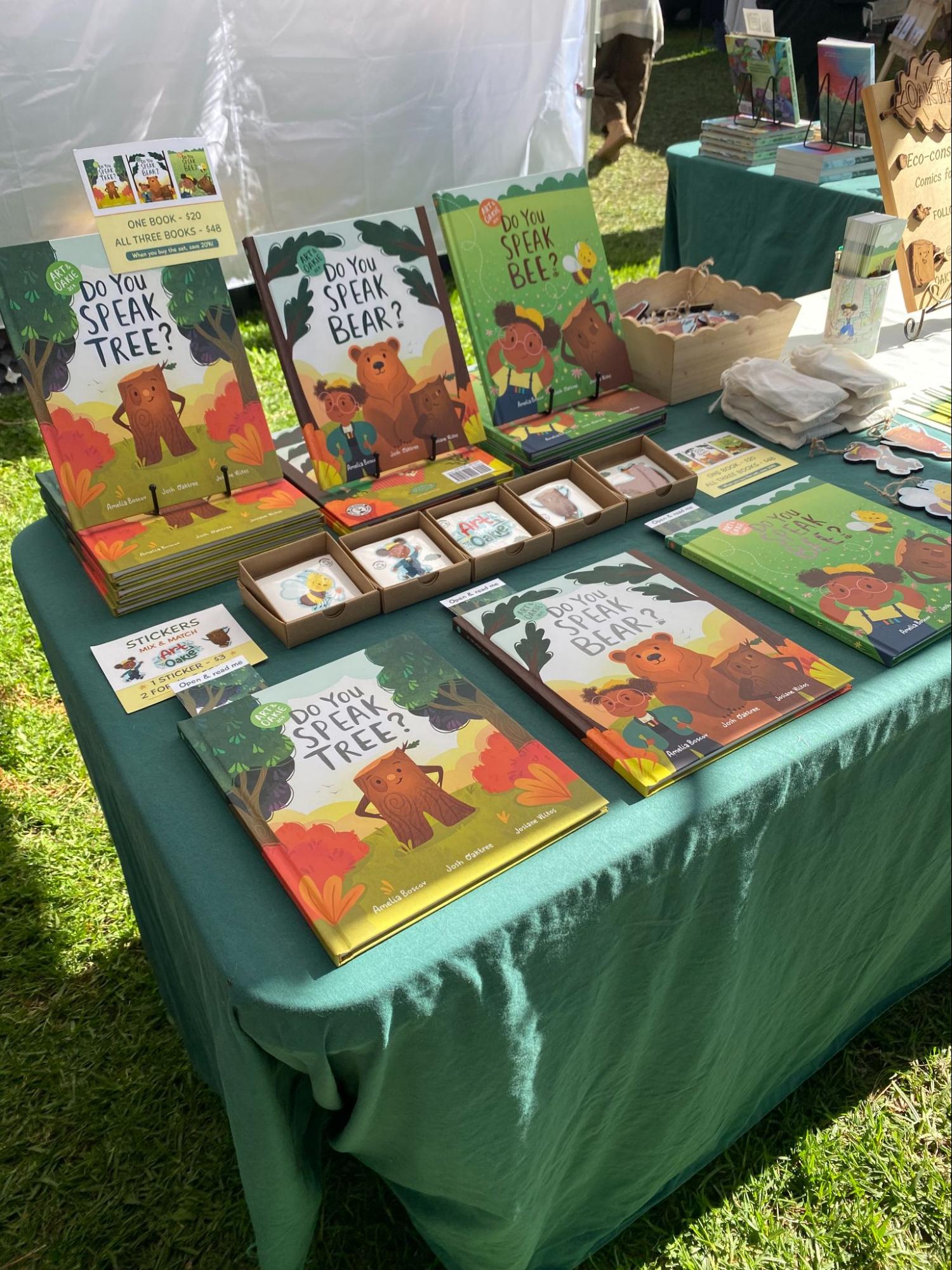 An outdoor table neatly displaying copies of Do You Speak Tree?, Do You Speak Bear?, and Do You Speak Bee?, with stickers and bookmarks arranged alongside