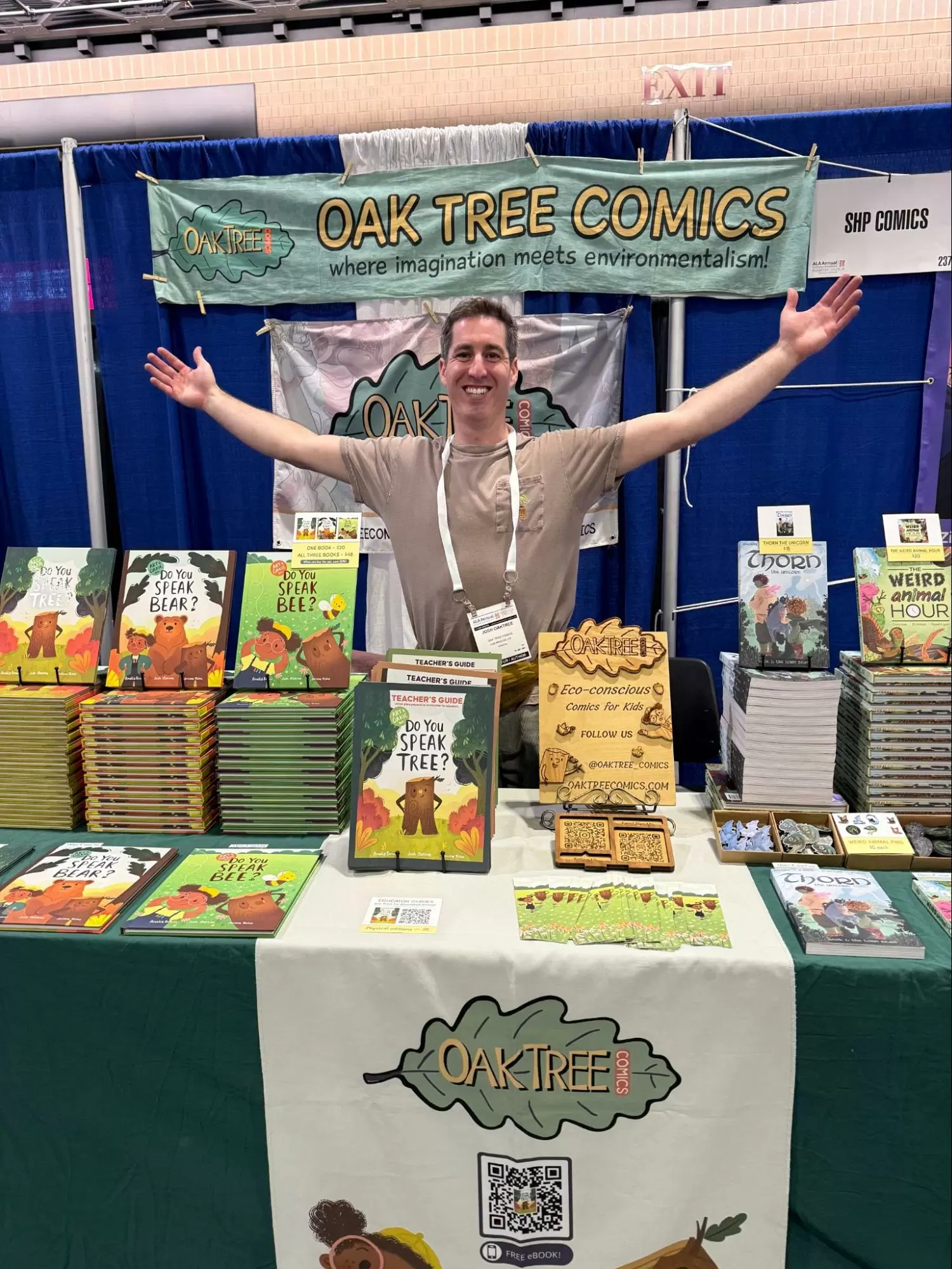 Josh Oaktree standing with his arms raised wide behind his Oak Tree Comics booth at an indoor comics convention, beneath a large hanging Oak Tree Comics banner, with tables full of his picture books