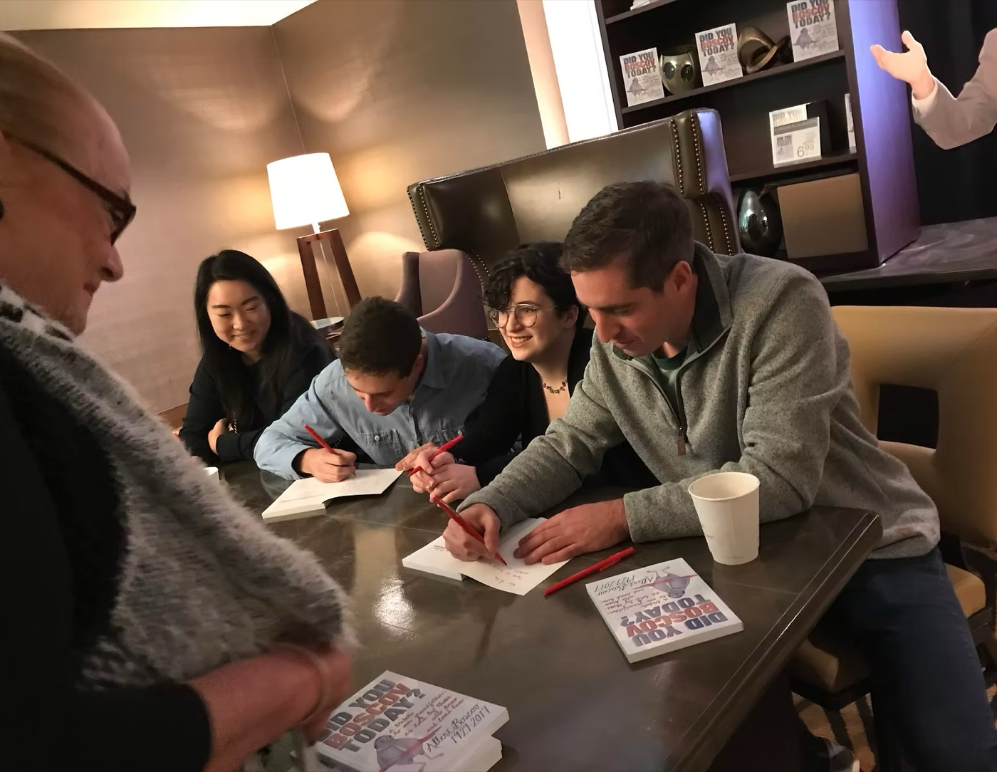 Photograph of Josh and his cousins seated at a table signing copies of Did You Boscov Today? during a book event at a Boscov's department store, with display copies of the book on the shelves behind them