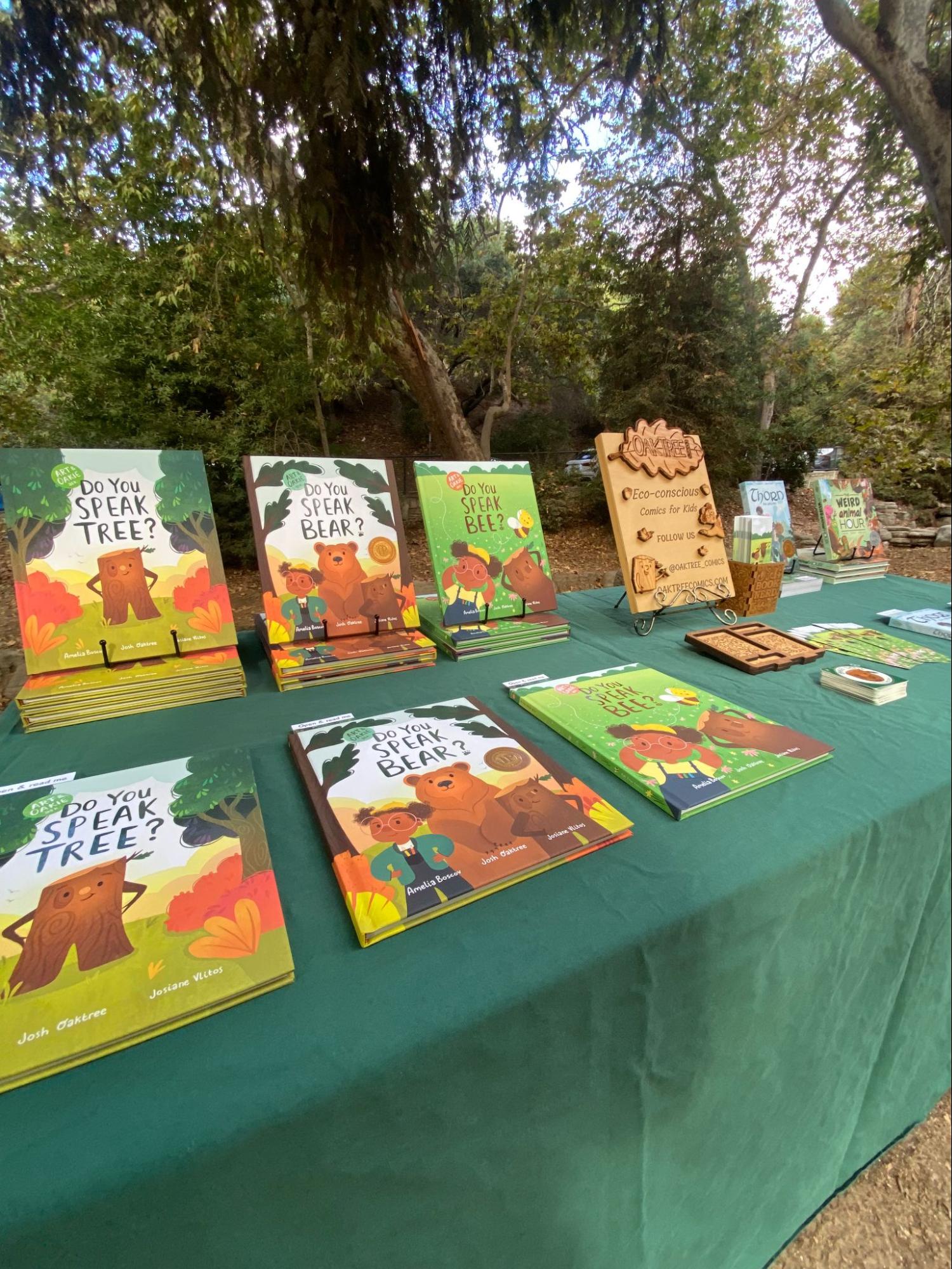 An outdoor table displaying copies of the Art and Oakie Ask picture books Do You Speak Tree?, Do You Speak Bear?, and Do You Speak Bee? standing upright and laid flat on a green tablecloth