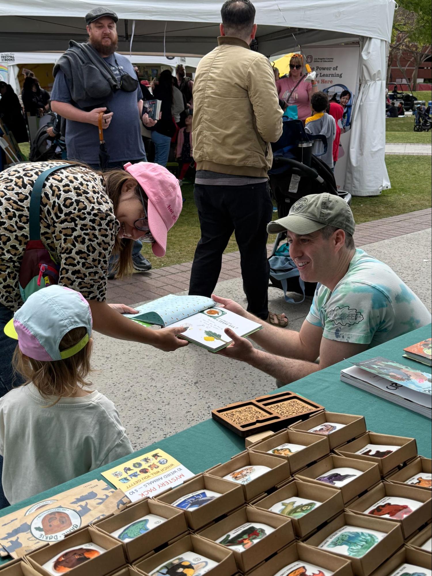 A child in a pink hat reaching across the booth table to take a picture book from a smiling Josh Oaktree at a busy outdoor festival