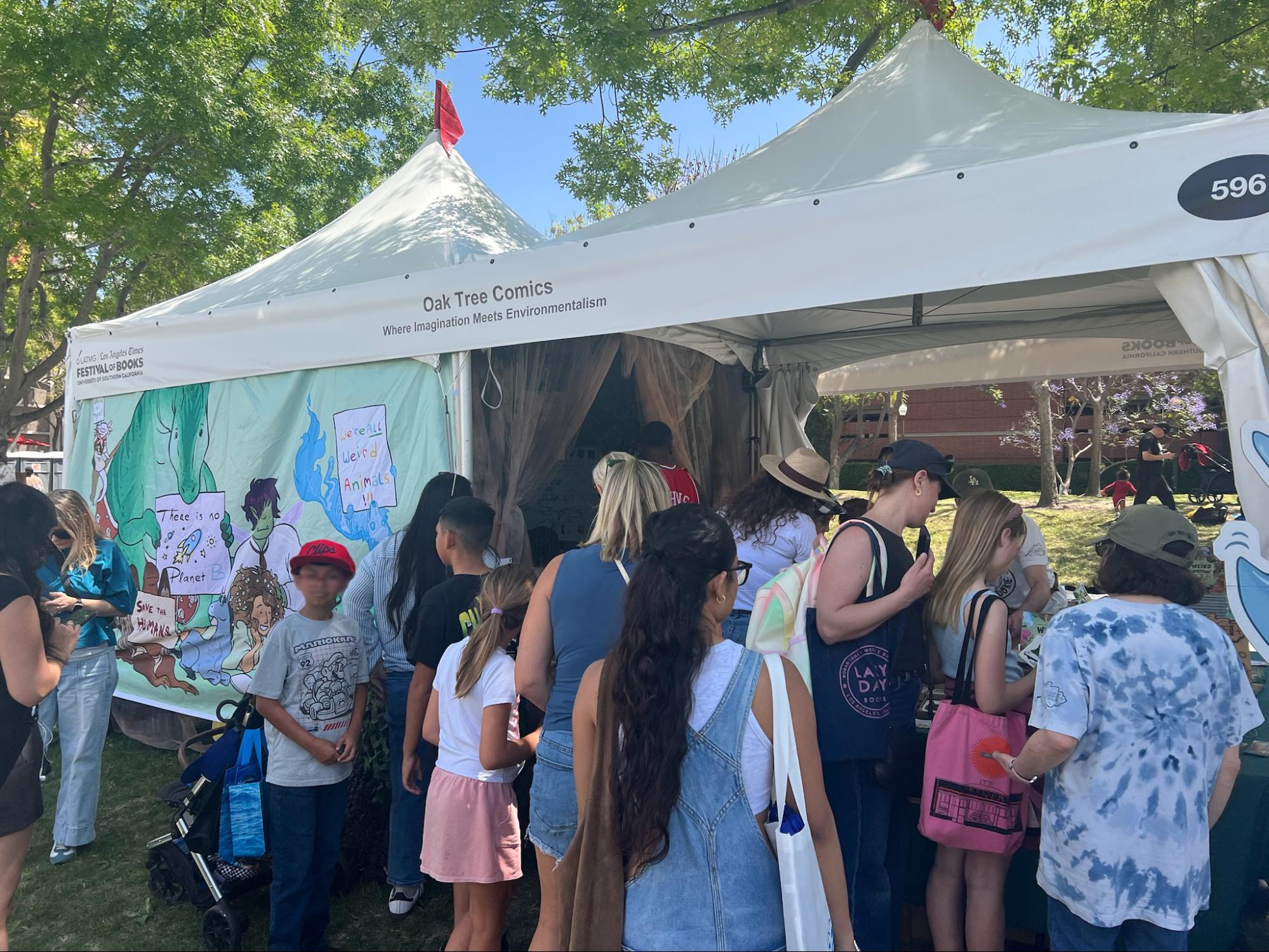 A crowd of festival-goers gathered at the Oak Tree Comics tent, browsing books in front of a hand-painted backdrop on a sunny day