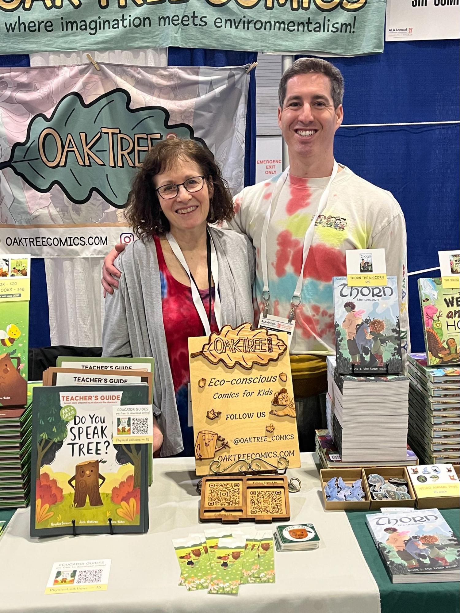 Josh Oaktree and his mother standing arm-in-arm and smiling behind the Oak Tree Comics booth at the American Library Association conference