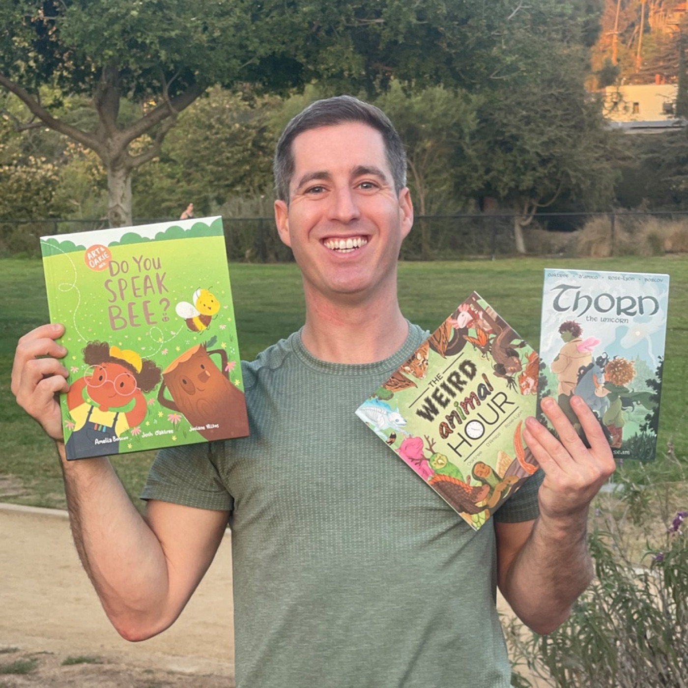 Josh Oaktree standing outdoors holding three Oak Tree Comics books, Do You Speak Bee?, The Weird Animal Hour, and Thorn the Unicorn