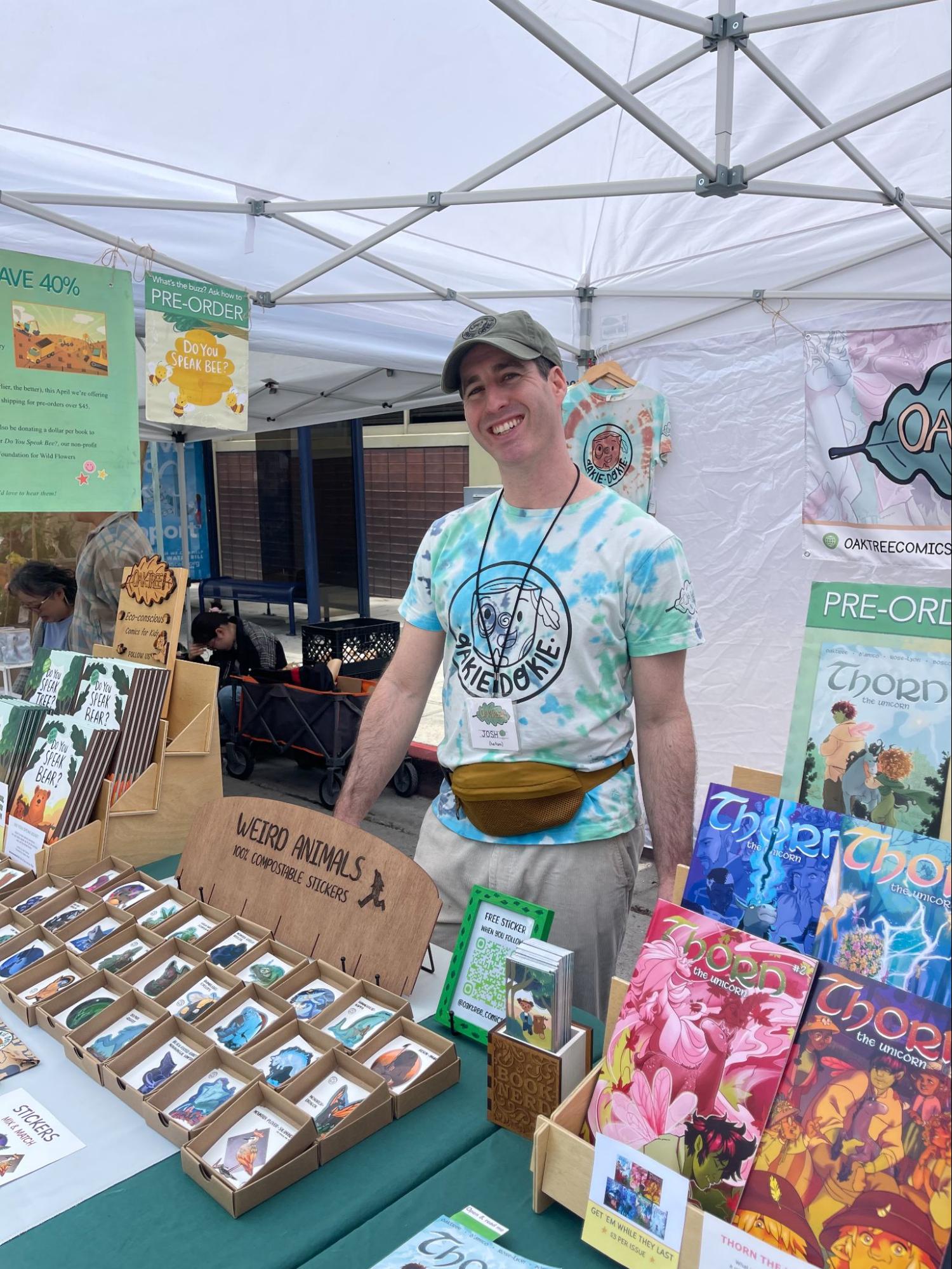 Josh Oaktree smiling behind his outdoor tent booth, surrounded by displays of his books and pre-order signage for upcoming titles
