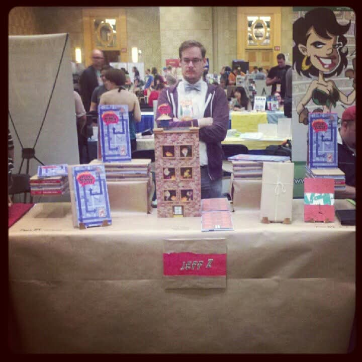Jeff's brother Adam in a blue bowtie and purple hoodie standing behind the 'Jeff Z' exhibitor table, which is stacked with copies of the book and a tall building-shaped cardboard display