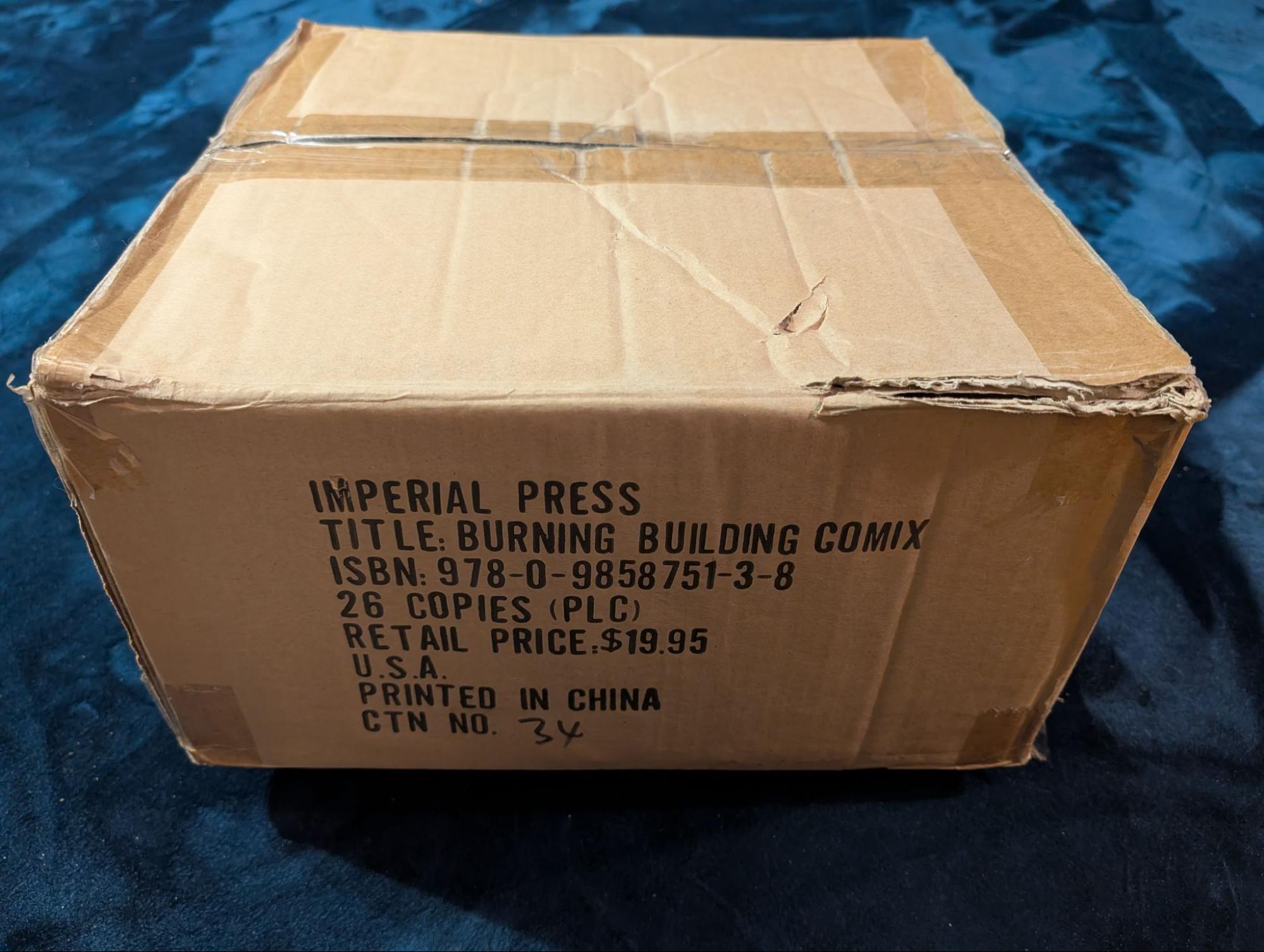 A worn cardboard shipping carton stamped 'Imperial Press, Title: Burning Building Comix, ISBN 978-0-9858751-3-8, 26 copies, retail price $19.95, U.S.A., Printed in China, CTN No. 34'