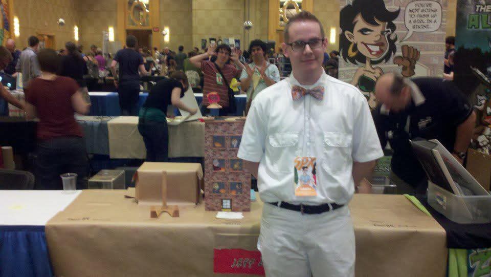 Jeff Zwirek in a white shirt and orange bowtie standing in front of his exhibitor table at SPX, with a busy convention hall full of attendees behind him