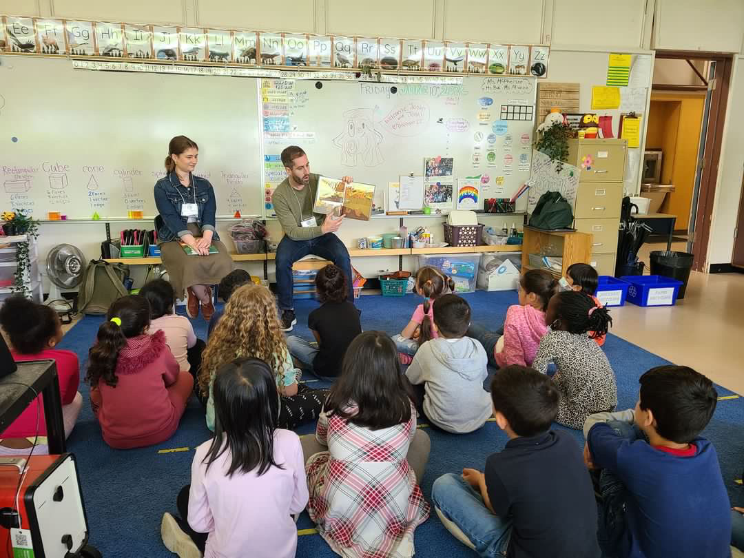 Josh Oaktree and a teacher seated at the front of a classroom, reading an illustrated book to a large group of young students seated on the floor