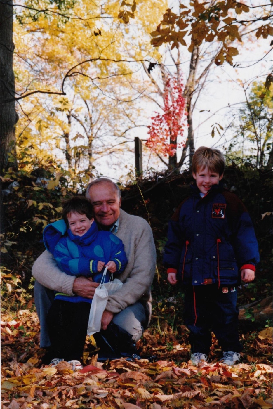 Young Josh as a small child being held by his grandfather Albert Boscov in a yard full of autumn leaves, with Josh's older brother standing nearby