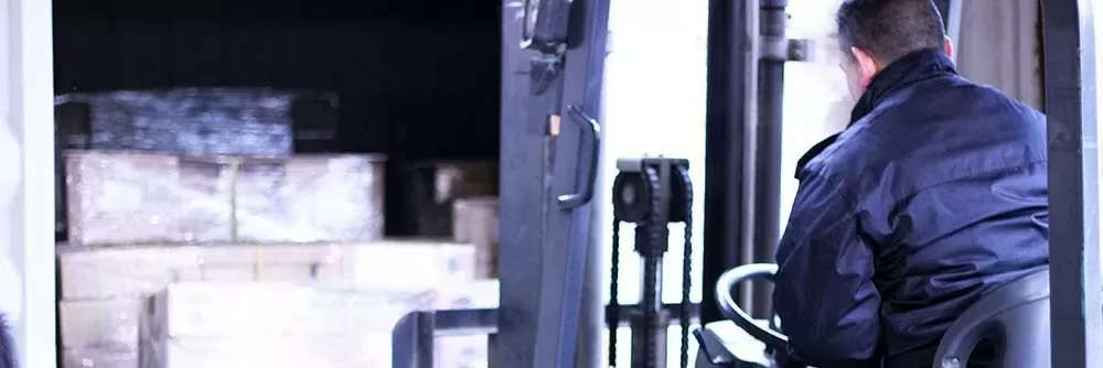 A forklift operator in a dark jacket driving pallets of cartons into a dimly lit container or trailer