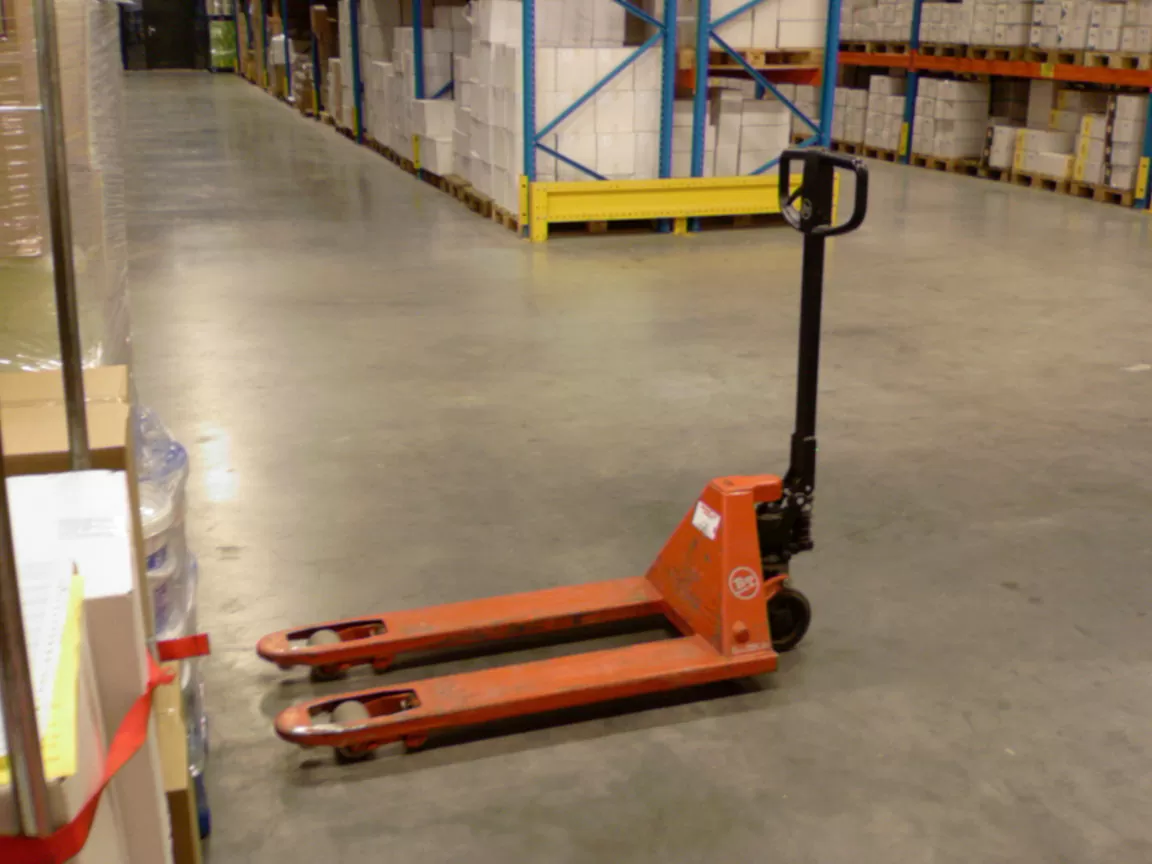 An orange pallet jack parked in a warehouse aisle beside racked and shrink-wrapped pallets