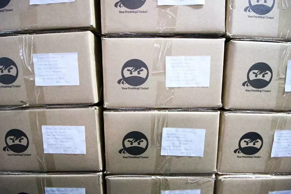 Stacked cartons printed with the PrintNinja ninja logo and the words Your PrintNinja Order, each shrink-wrapped and carrying a printed shipping label