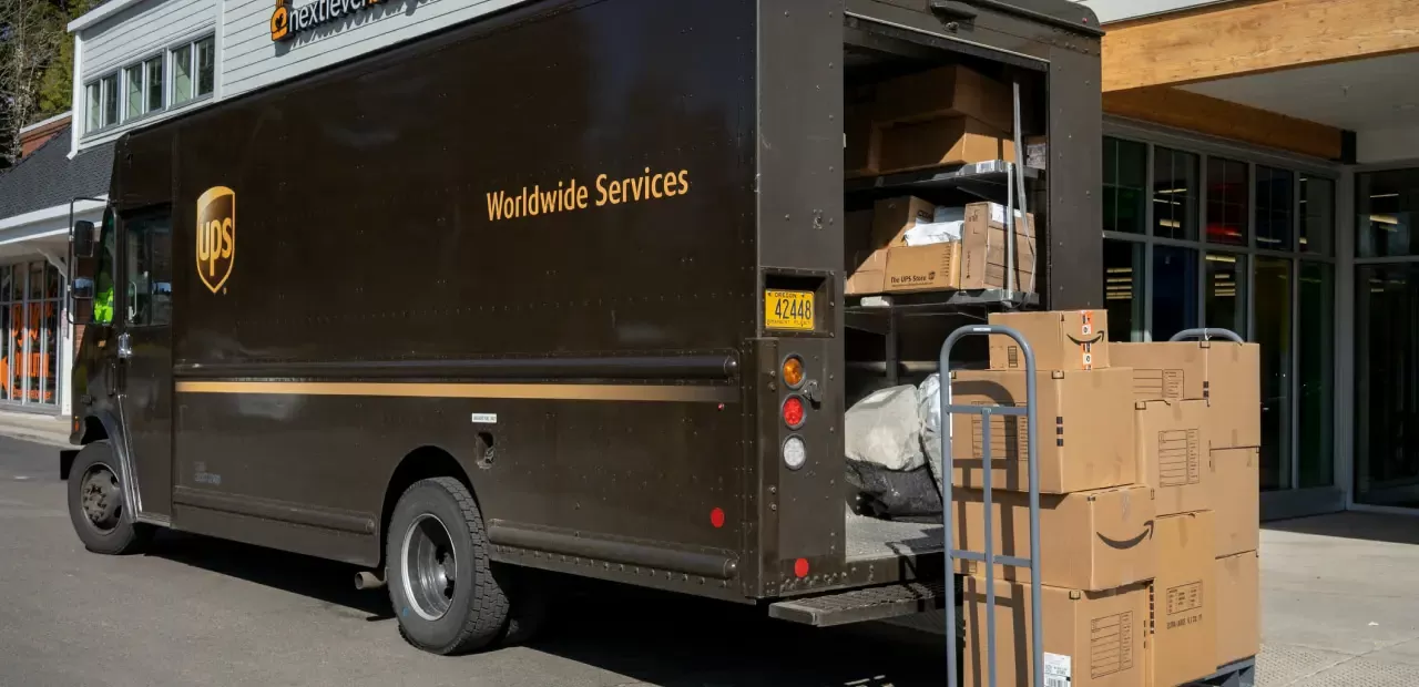 A brown UPS delivery truck parked with its rear door open, a hand cart stacked with cardboard boxes standing beside it on the sidewalk